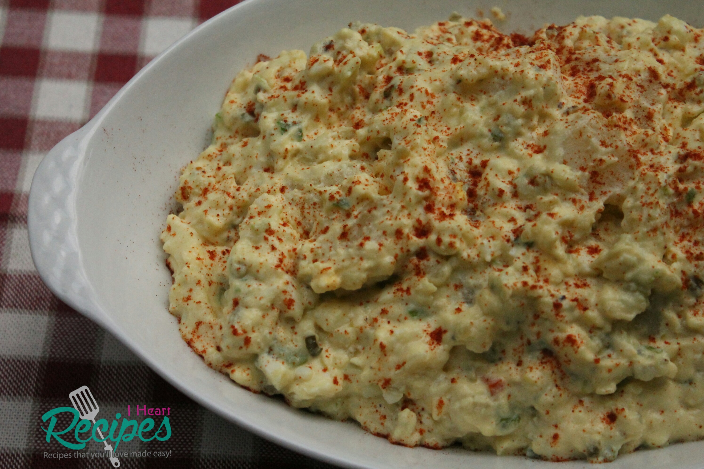 An overhead image of a Southern-style potato salad served in a white casserole dish. The dish sits on a red and white checkered tablecloth.