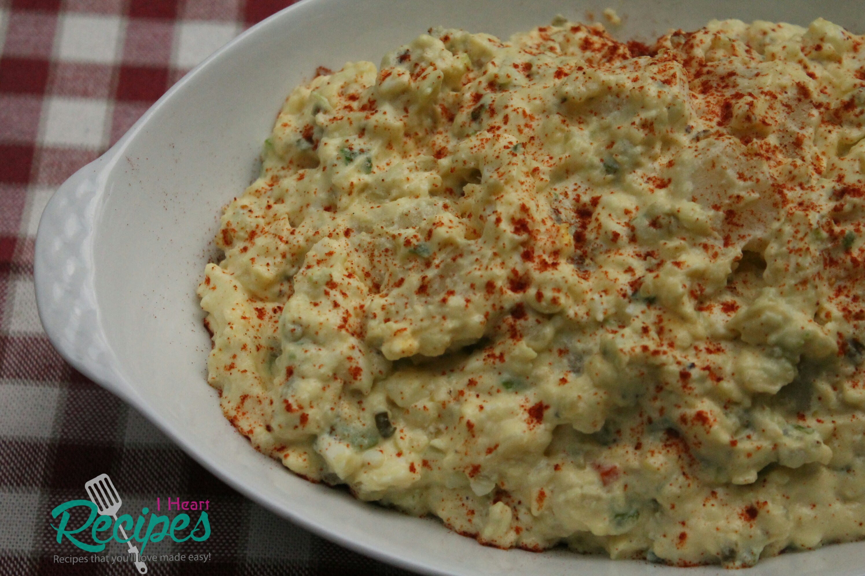A round, white casserole dish filled with creamy Southern potato salad. The potato salad dressing is a light yellow color, mixed with chunky potatoes and vegetables and sprinkled with paprika.