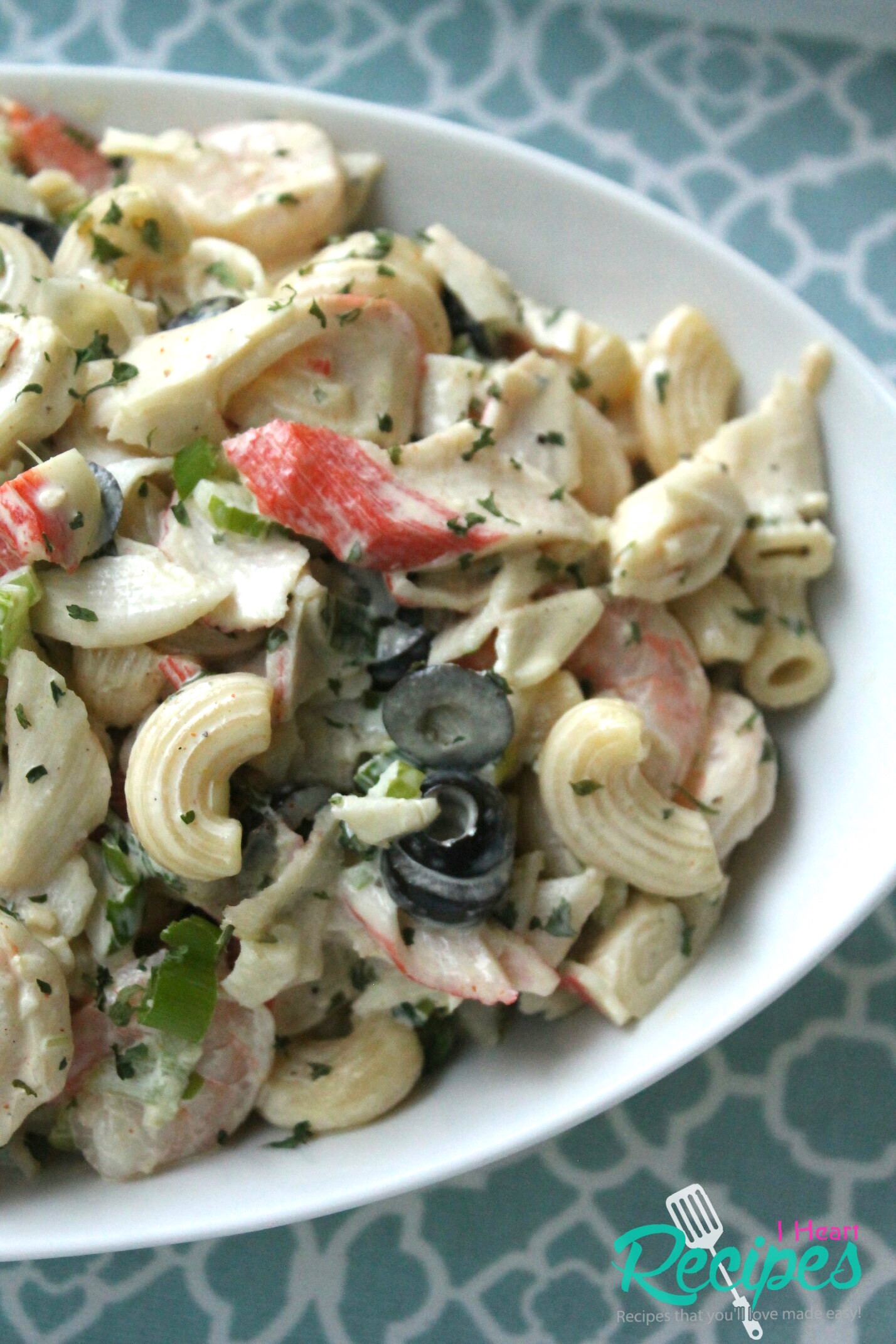 A close-up image of a large bowl of seafood macaroni salad, made with crab, shrimp, macaroni pasta, olives, celery, and onions mixed with a light and creamy dressing.