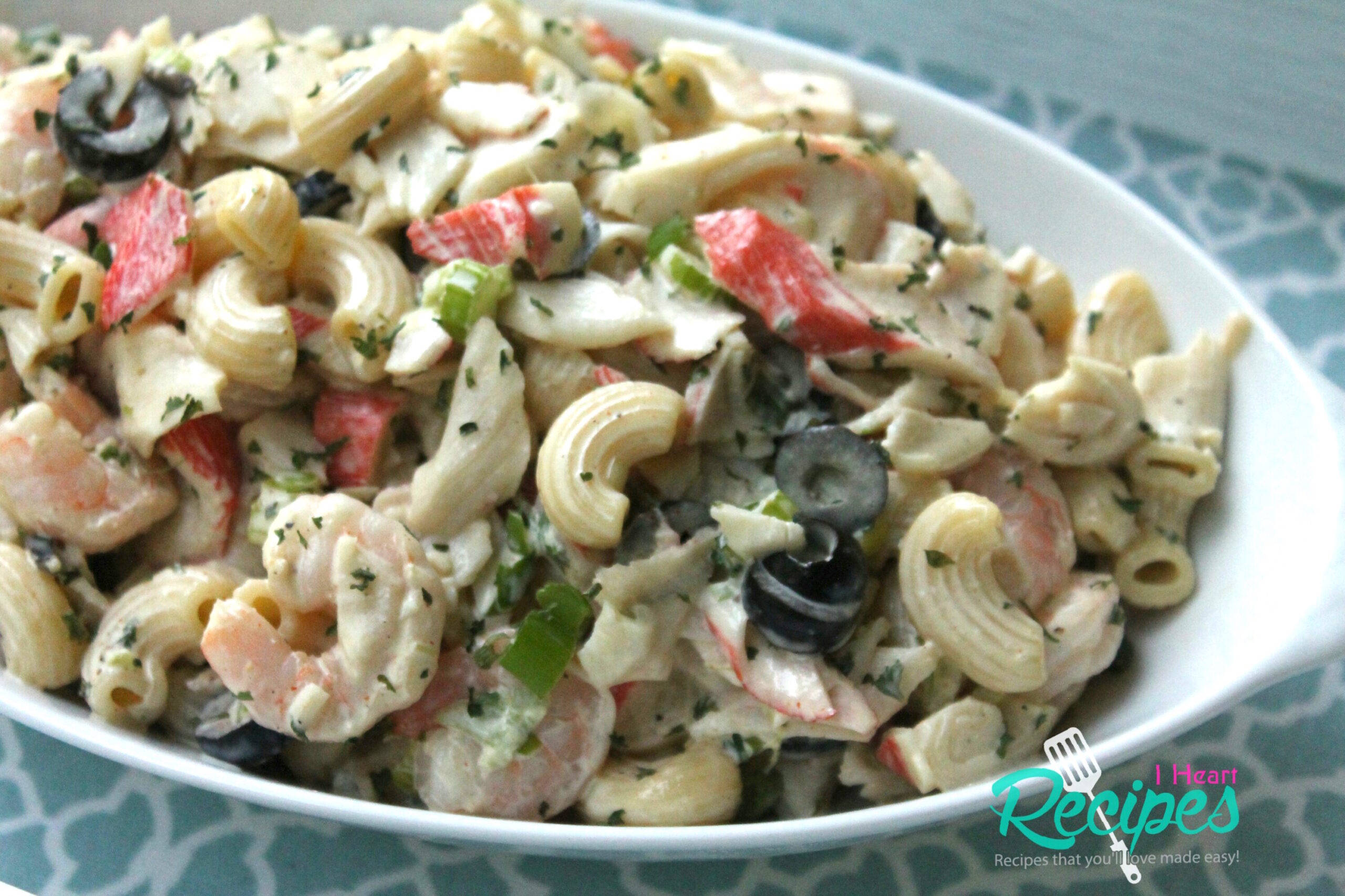 A large white, oval dish is filled with creamy seafood salad. Chunks of crab meat, shrimp, pasta, and veggies are mixed with a creamy mayonnaise dressing.
