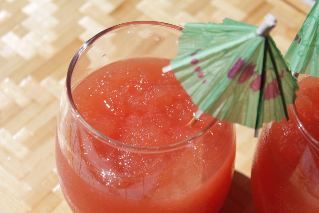 An sunny overhead shot of bright red watermelon vodka slushies garnished with a green paper cocktail umbrella. 