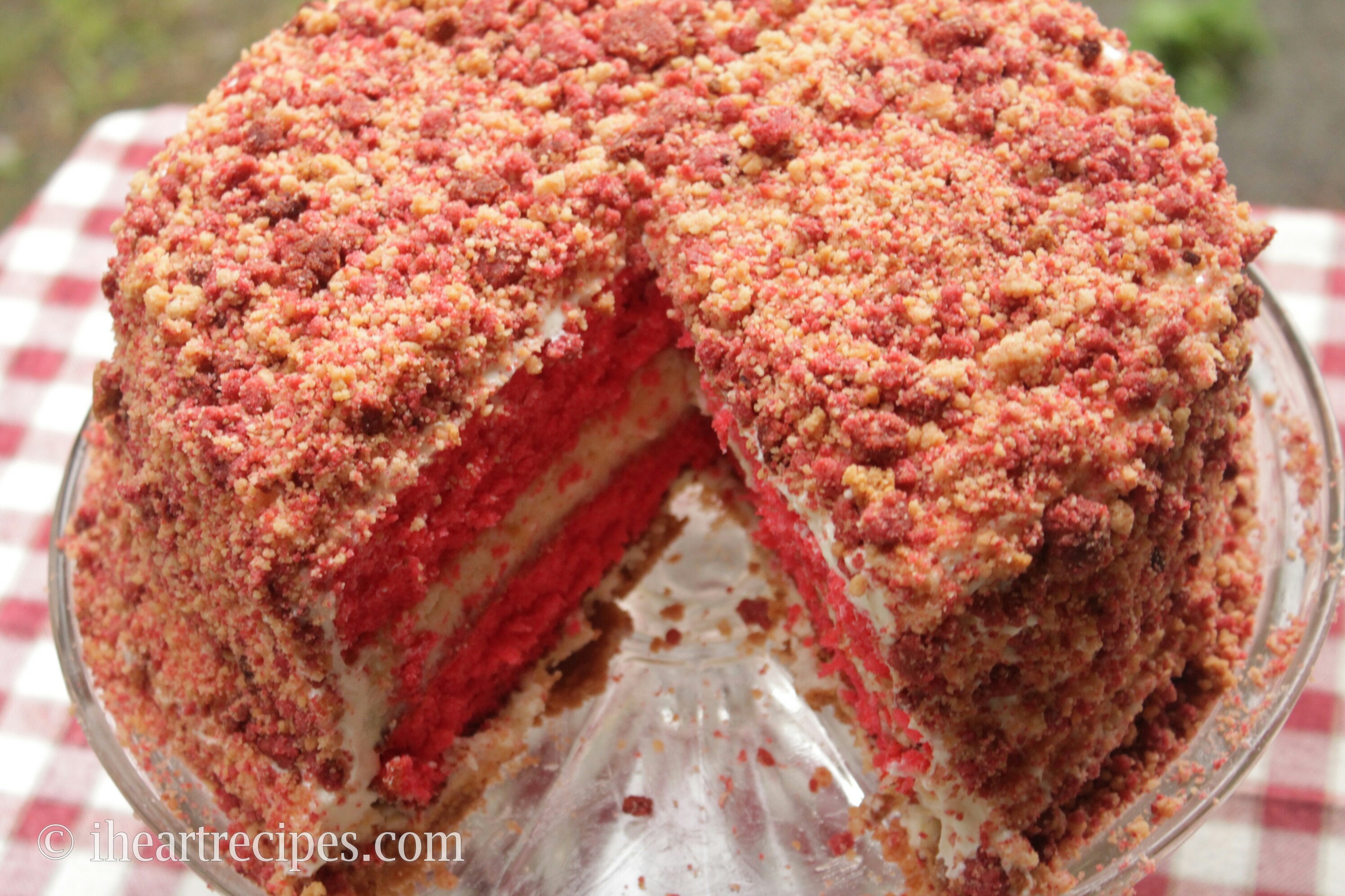 An overhead view of the strawberry shortcake cake, showing the crunchy strawberry cookie coating around the entire cake. A single slice is cut out, revealing the layers of cheesecake and strawberry cake inside.