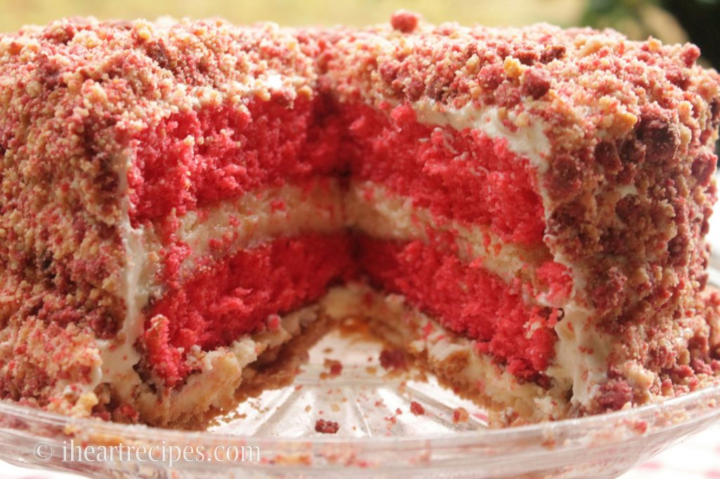 A whole strawberry shortcake cheesecake, topped with a strawberry cookie crumble topping, with a large slice cut out, showing the four layers of strawberry cake and cheesecake inside.