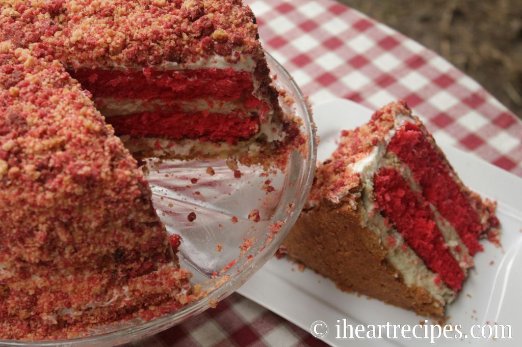 A strawberry shortcake cheesecake with a single slice cut out is served on a white plate, made with layers of strawberry cake, vanilla cheesecake, and vanilla cookie crumble.