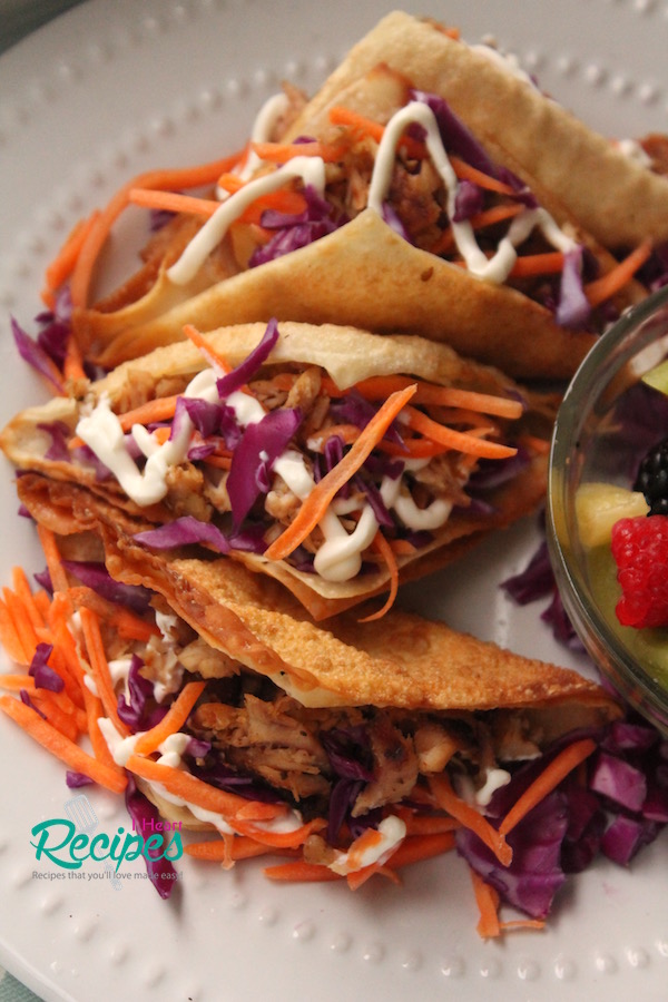 A row of chicken wonton tacos served on a white plate next to a glass bowl full of fresh fruit. 