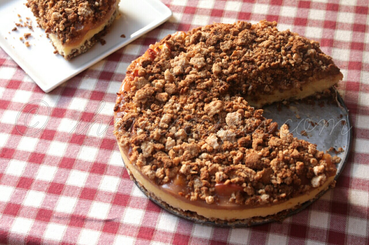 A whole peach cobbler cheesecake served on a metal pan, with a single slice cut out and served on a small plate. The cheesecake sits on a table covered with a red and white checkered tablecloth.