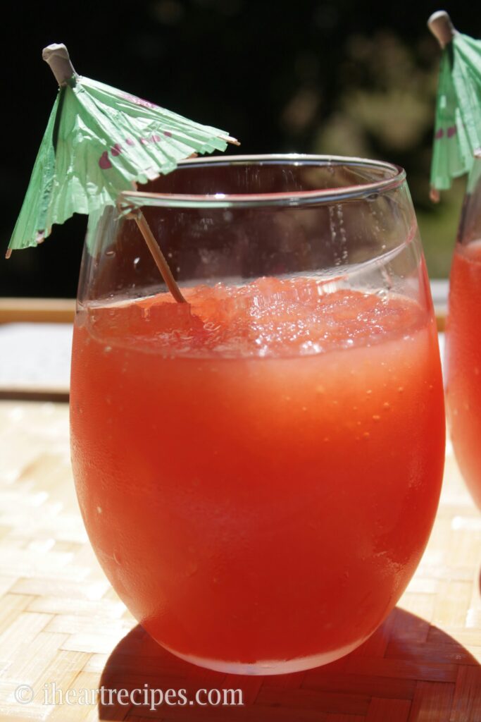 A clear glasses filled with bright red homemade watermelon vodka slushies and garnished with a paper umbrella. 