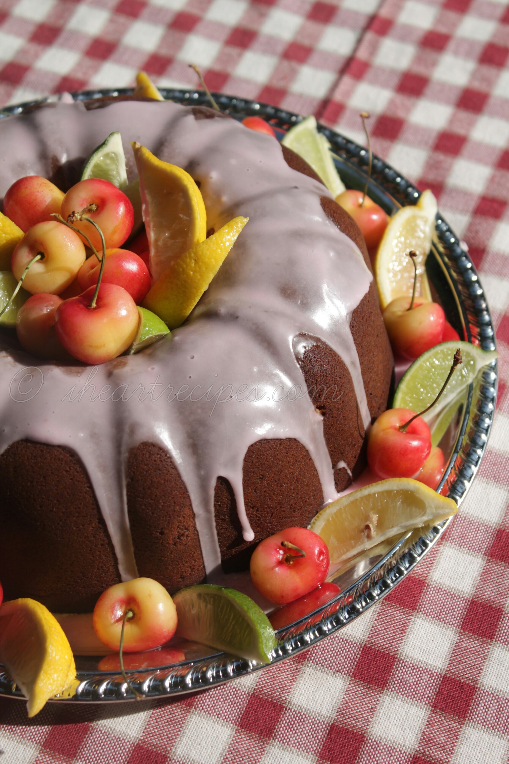 A perfect bundt cake with sweet vanilla icing garnished with fresh fruit slices served on a scalloped metal tray set on a red and white checkered tablecloth.