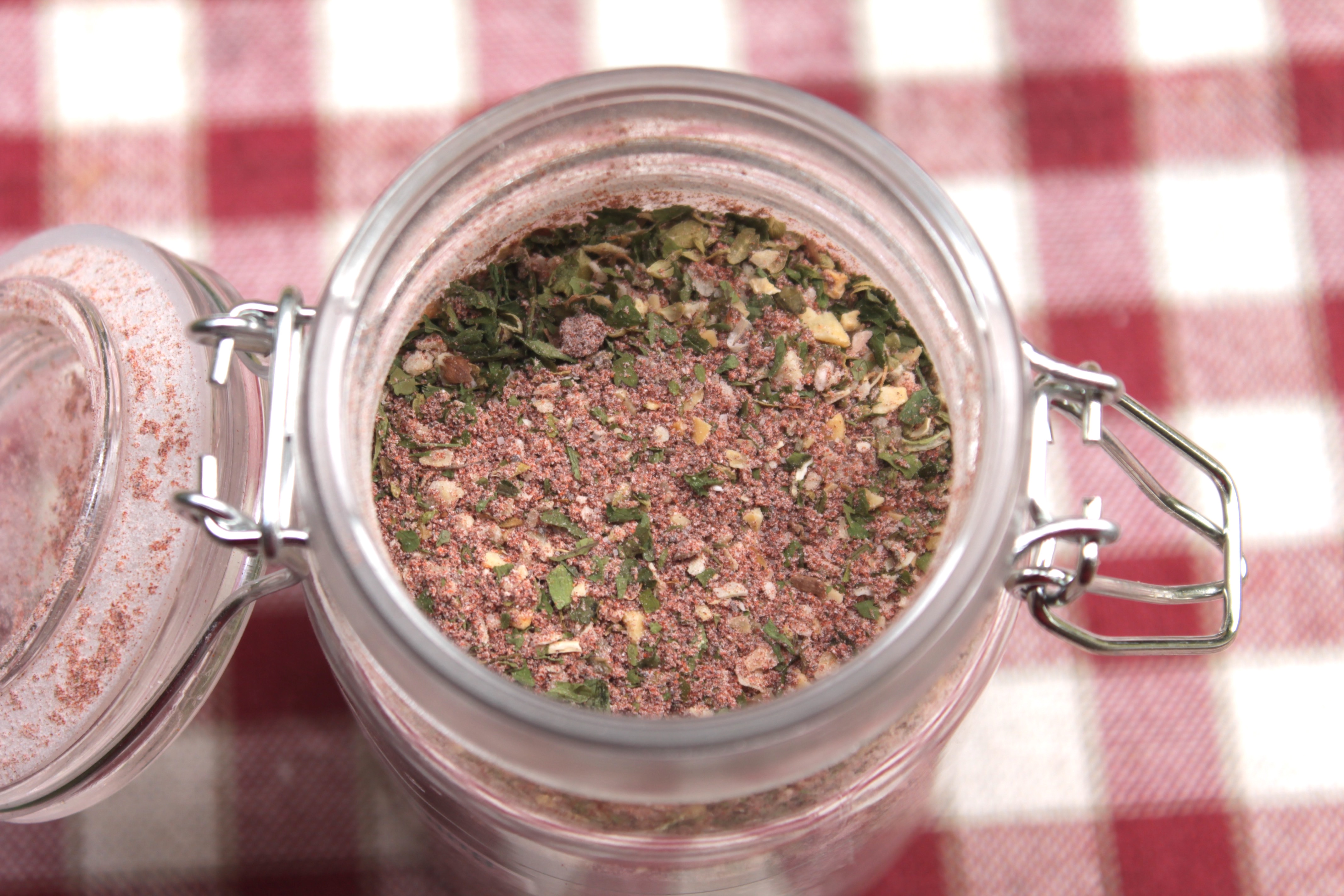 Homemade taco seasoning fills a glass jar sitting on a red and white checked tablecloth. 