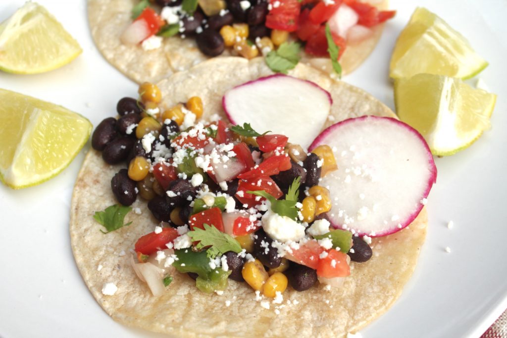 Vegetables and beans on two corn tortillas garnished with radishes and pieces of lime served on a white plate. 