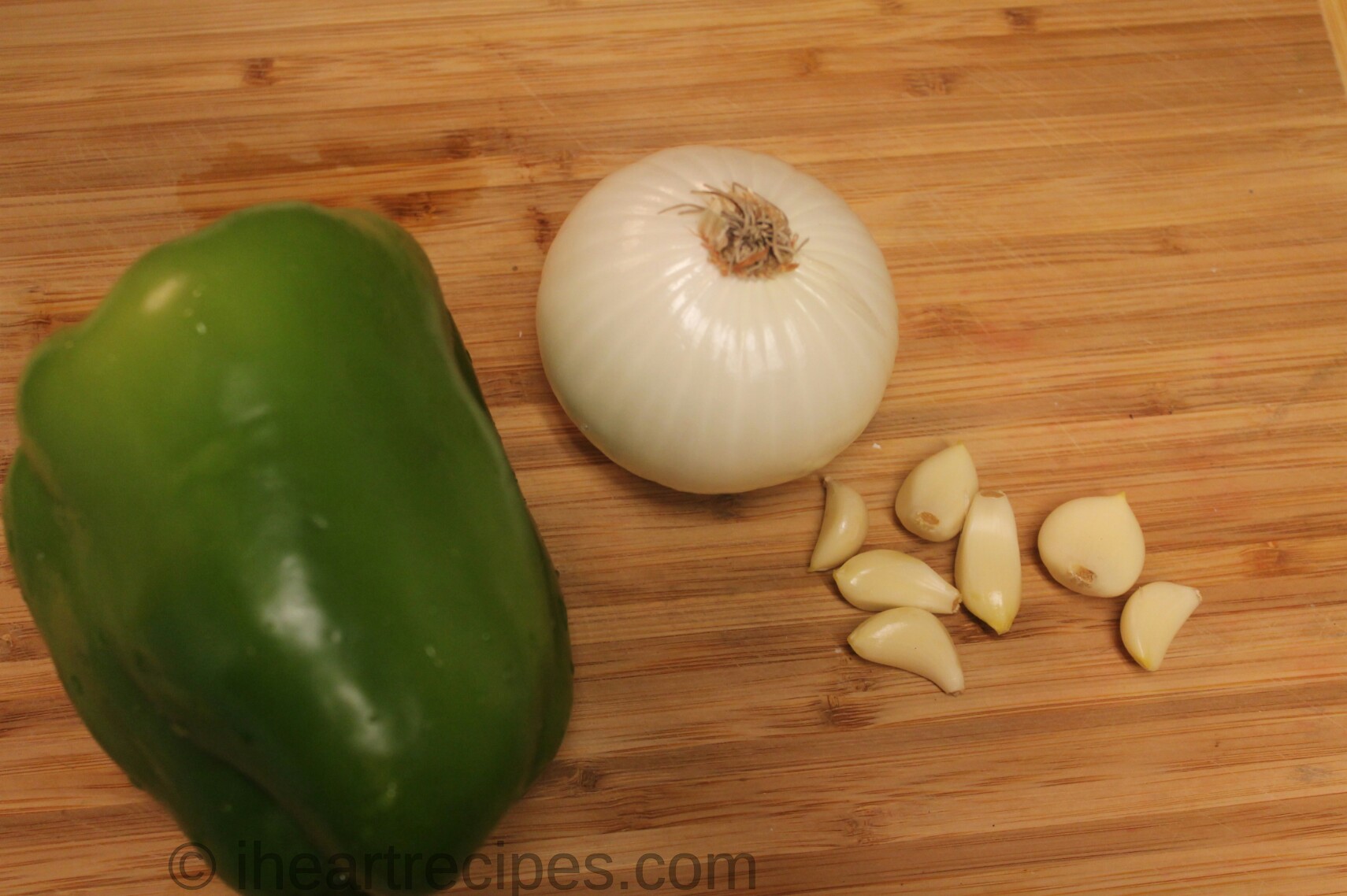 A large green bell pepper, white onion, and several fresh garlic cloves sit on a wooden cutting board.