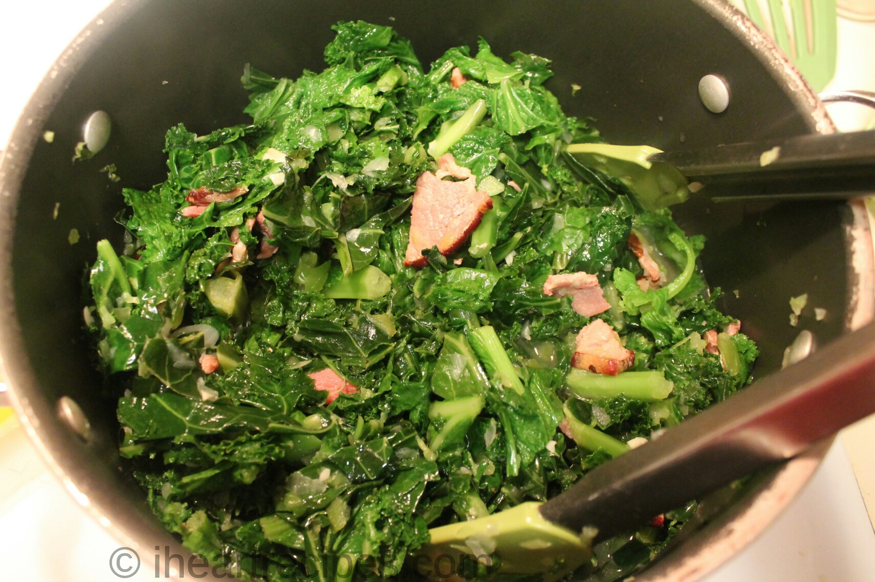 Mixed greens cook down in a large pot, mixed with cooked bacon, onions, peppers, and garlic.