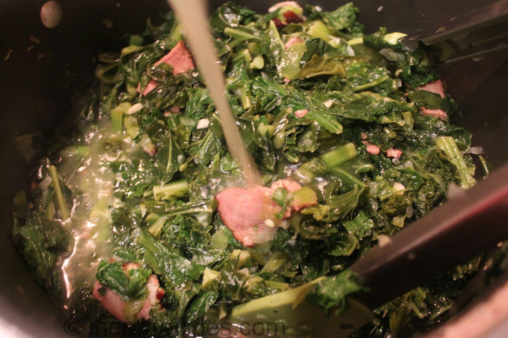 A person pours a steady stream of chicken broth into a pot of Southern mixed greens with vegetables and bacon.