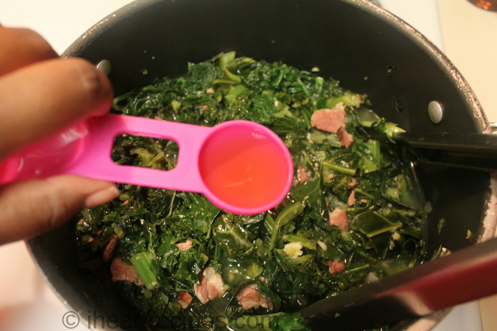 A person holds a tablespoon measuring spoon filled with apple cider vinegar over a large pot of Southern mixed greens.