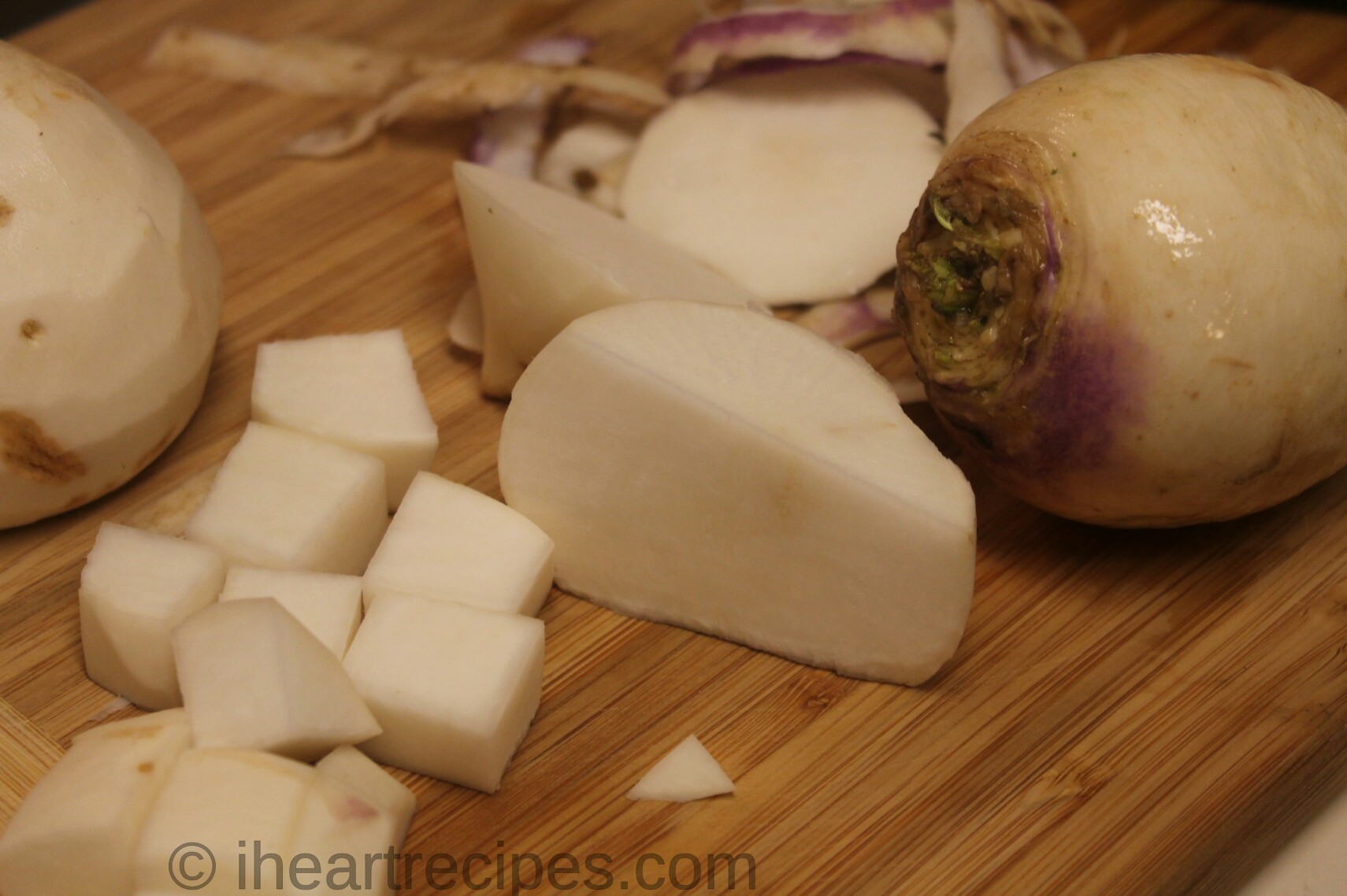 Raw turnips sit on a wooden cutting board. Some are chopped into cubes, some are cut in half, and one whole turnip remains unpeeled.