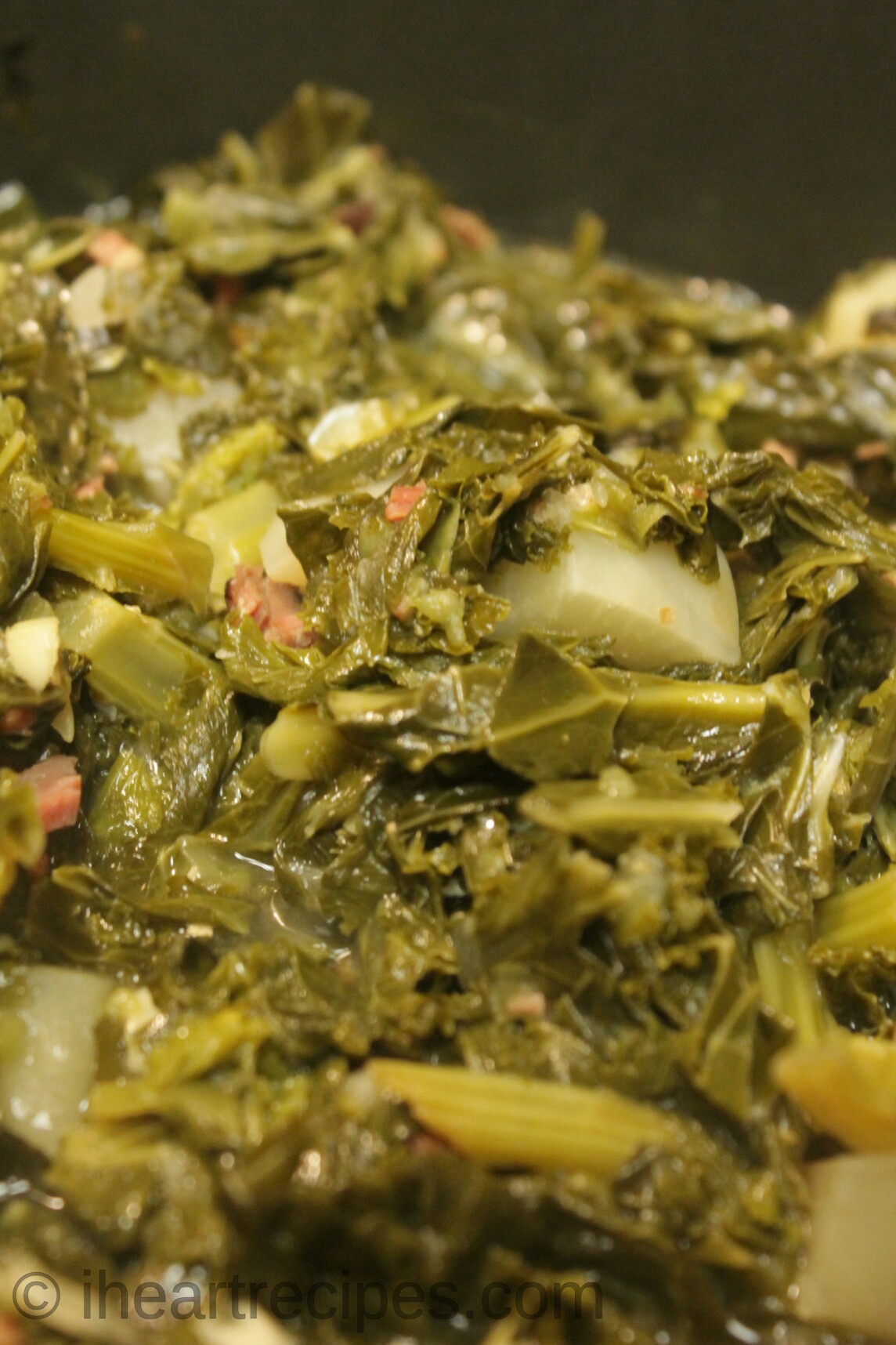 A close-up image of tender mixed greens cooked in a large pot with onions, bacon, bell peppers, garlic, and chicken broth.