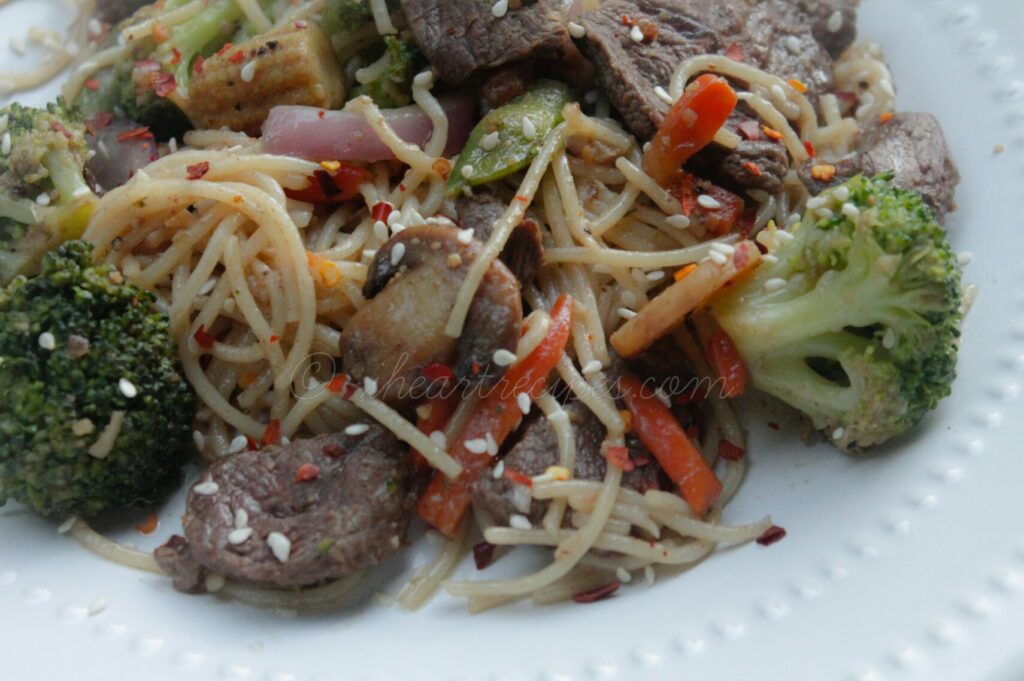 A healthy serving of beef and vegetable stir fry is on a white plate. The beef, vegetable, and noodle stir fry is garnished with sesame seeds and red pepper flakes.