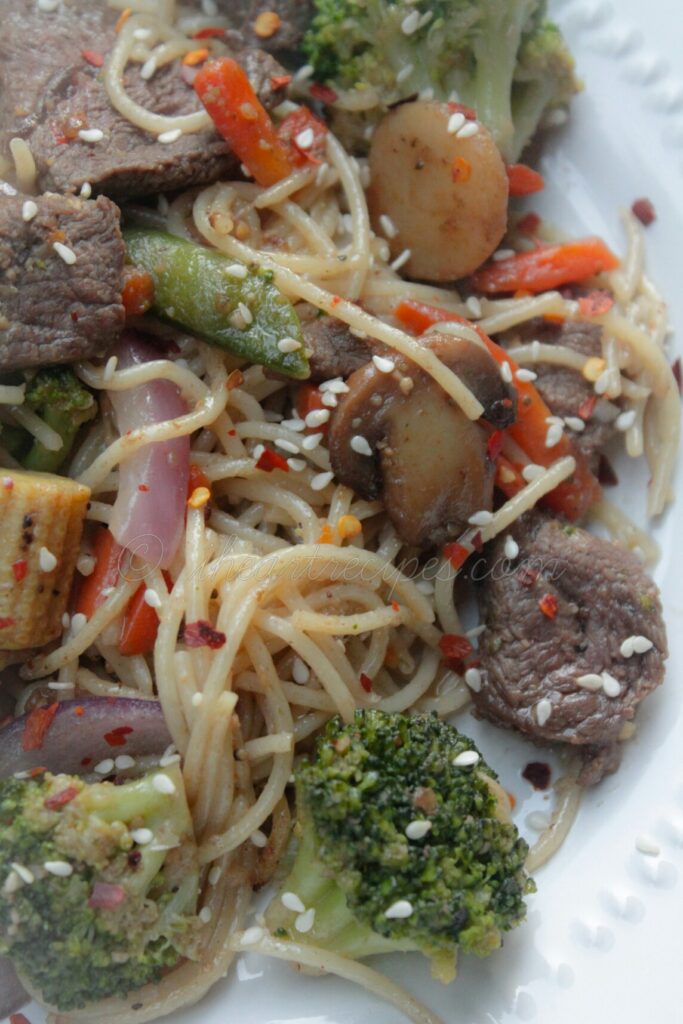 A close up image of beef and vegetable stir fry with noodles, cooked in an umami sauce and garnished with sesame seeds and red pepper flakes.