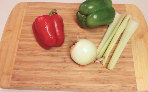 Fresh red and green peppers, yellow onion and stalks of celery on a wooden cutting board.