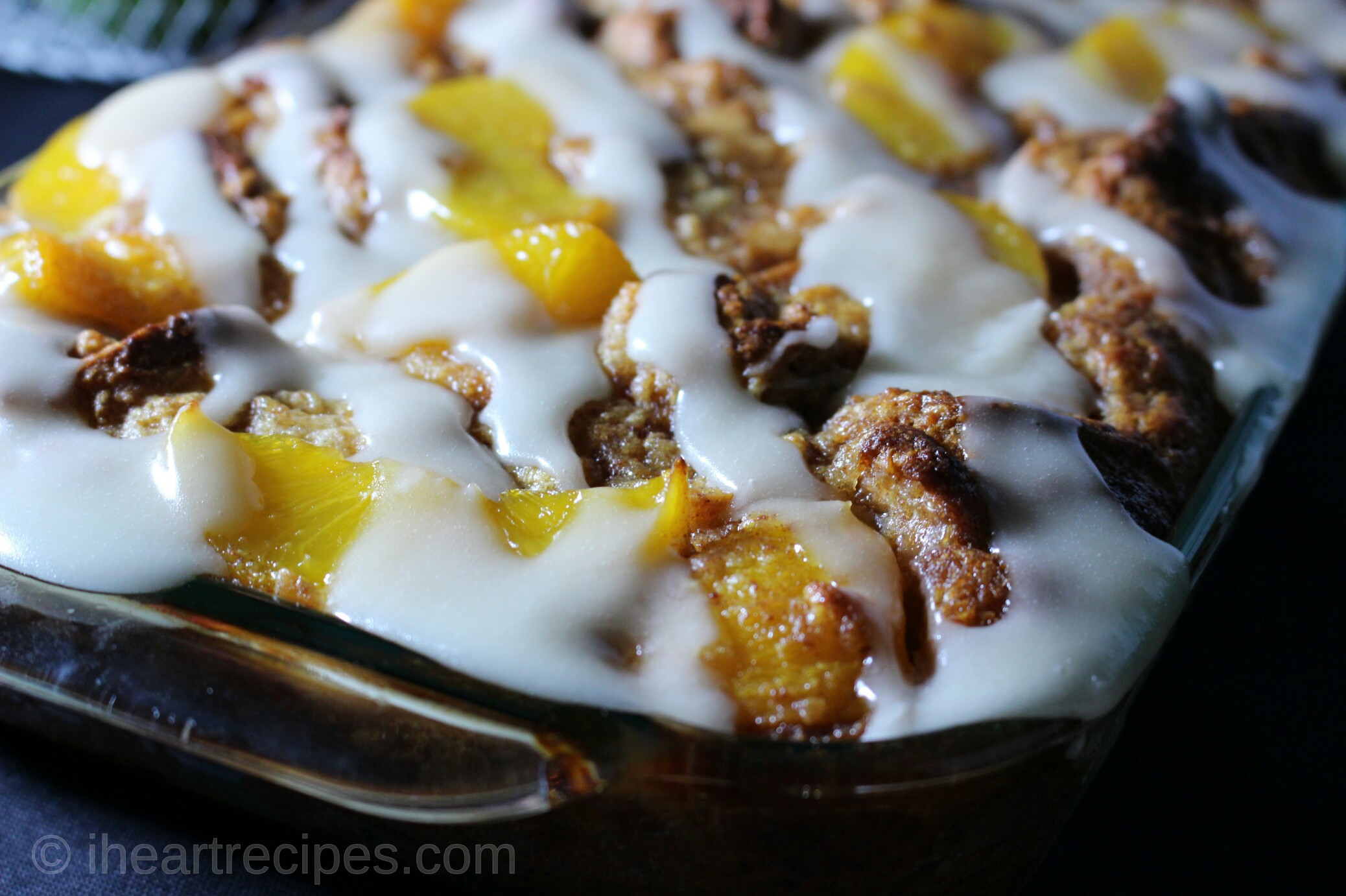A closeup image of a baking dish of peach cobbler bread pudding. The freshly baked dessert is topped with peach slices and a sweet vanilla icing.