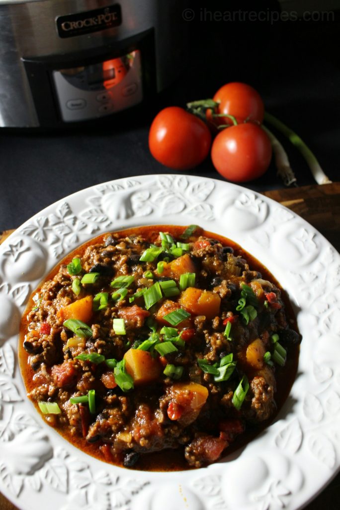 A bowlful of beef, black bean and butternut squash chili on a wooden table next to a black and silver Crock-Pot, three red vine tomatoes, and two green onions. 