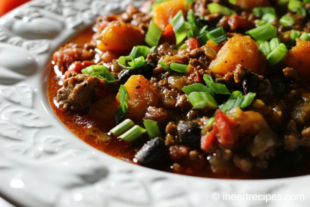 A close up image of beef, black bean and butternut squash chili garnished with green onions heaped into a decorative white bowl. 