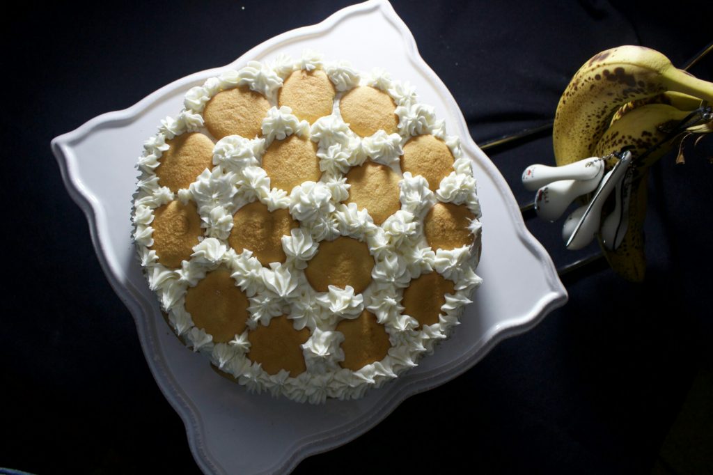 An overhead image of a semi-homemade banana pudding cake, decorated with dollops of vanilla frosting and vanilla wafer cookies. The cake sits on a square cake platter, next to a bunch of ripe bananas.