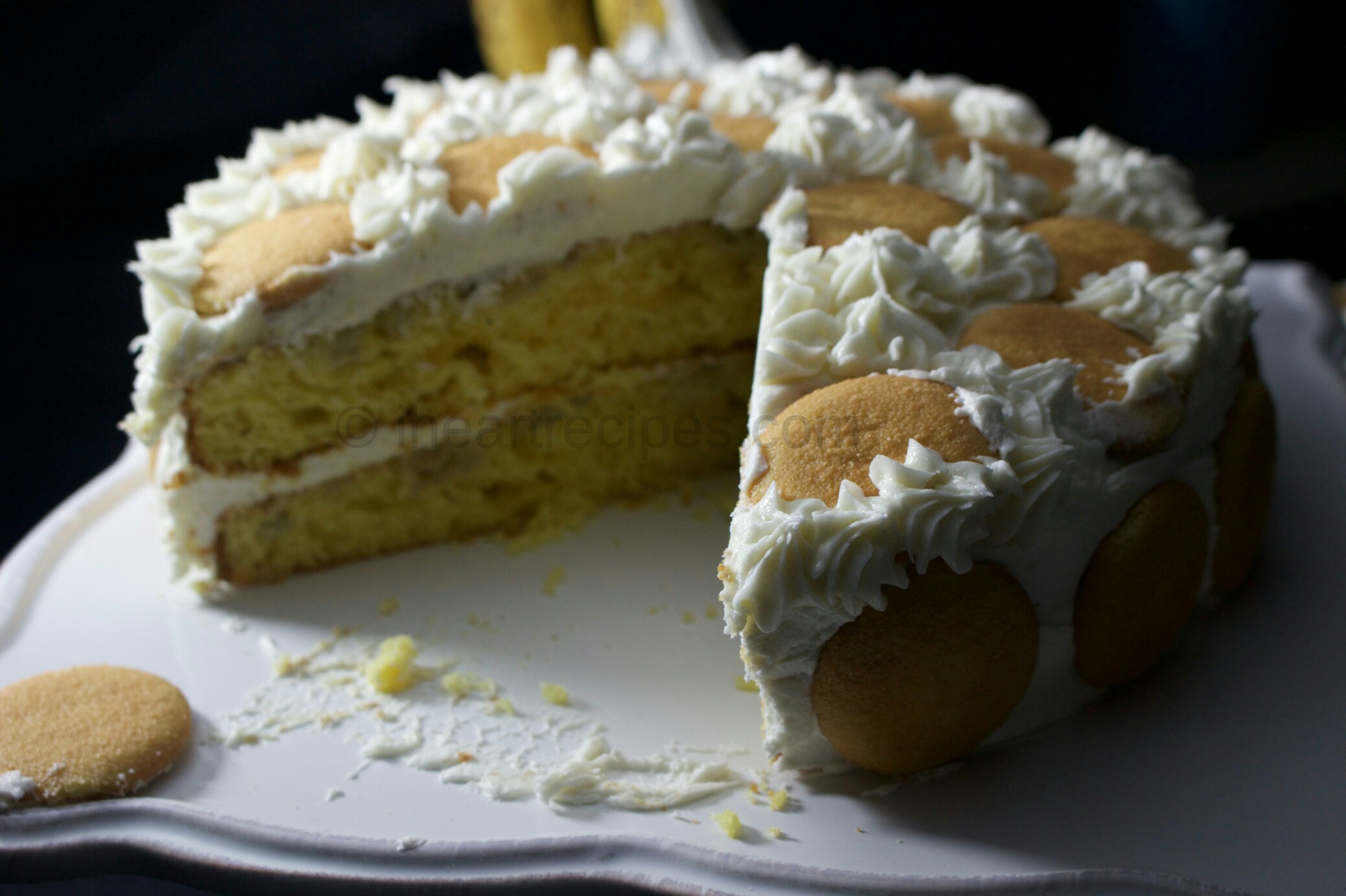 A side angle view of a two-layer banana pudding cake. The moist yellow banana cake is frosted with a homemade vanilla icing and topped with Nilla wafers. A large slice has been cut from the cake, showing the moist yellow cake layers inside.