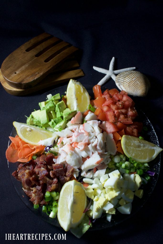 Tender crab, crispy bacon and colorful vegetables garnished with lemon wedges served on an elegant glass platter. Wooden salad forks and seashells lay nearby. 