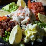 A colorful bowl of Crab Louie salad features eggs, carrots, tomatoes, avocado, crab, and lemon wedges.