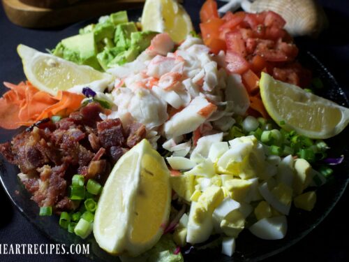 A colorful bowl of Crab Louie salad features eggs, carrots, tomatoes, avocado, crab, and lemon wedges.