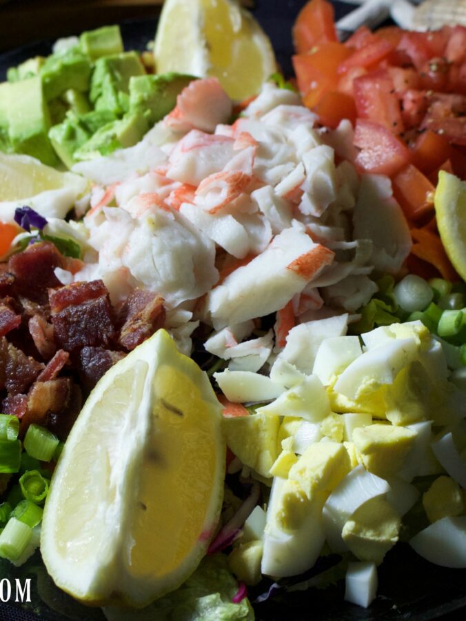 A colorful bowl of Crab Louie salad features eggs, carrots, tomatoes, avocado, crab, and lemon wedges.