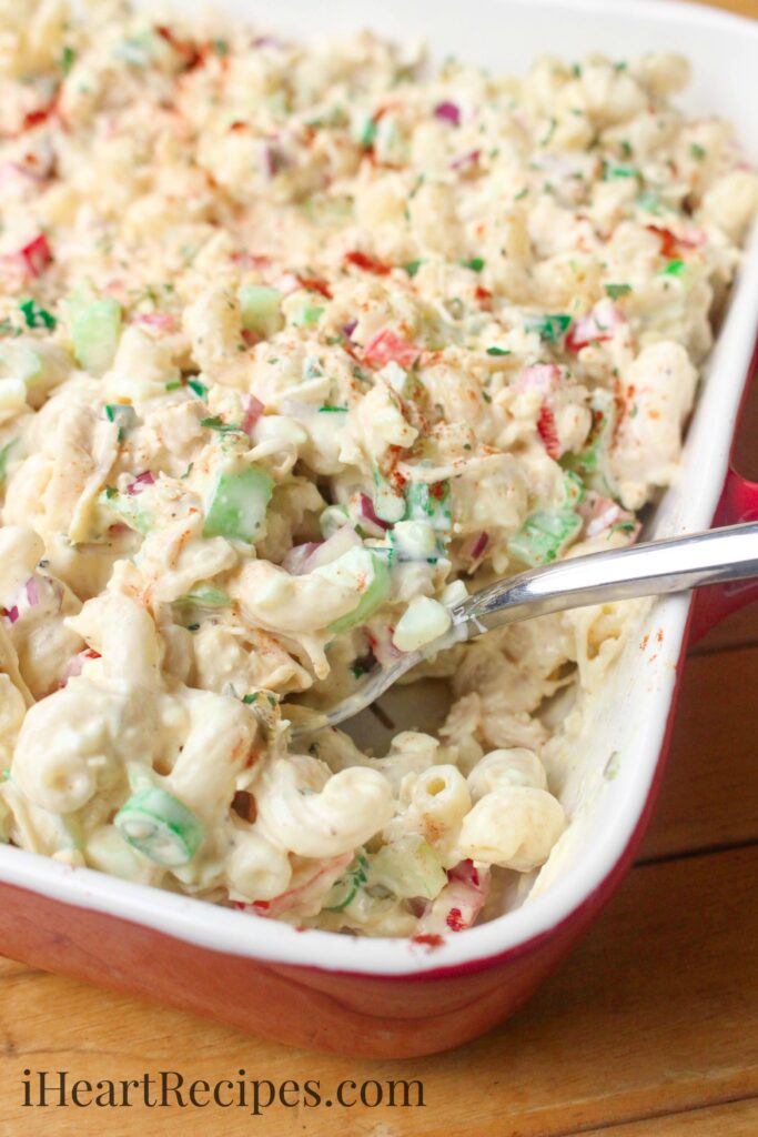 A white and red casserole dish is filled with a creamy macaroni salad with chicken, celery, red bell pepper, and other vegetables. A serving spoon holds a generous scoop of the creamy pasta salad.