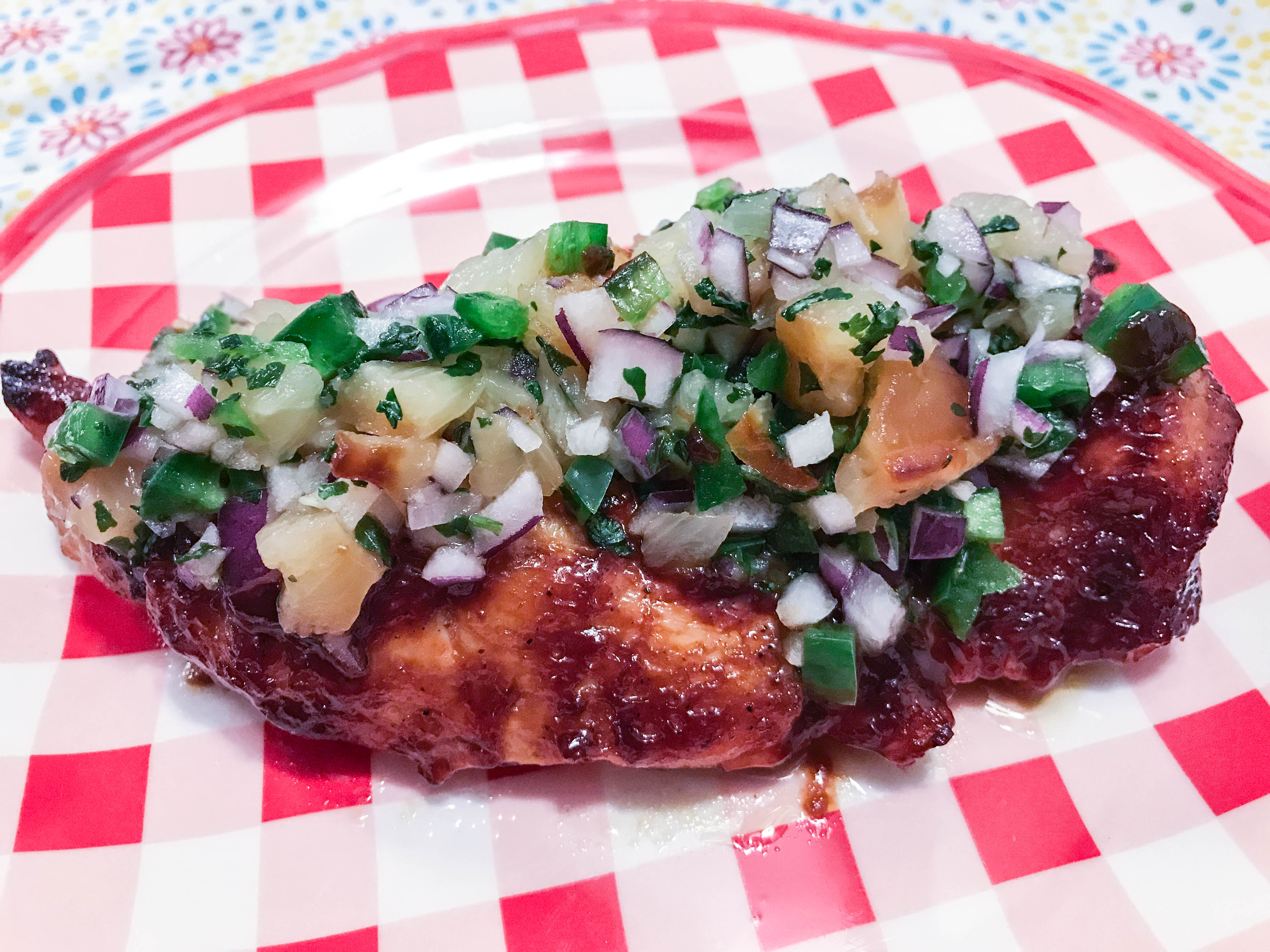 A sweet and spicy chicken breast topped with homemade pineapple salsa served on a red and white checked plate.