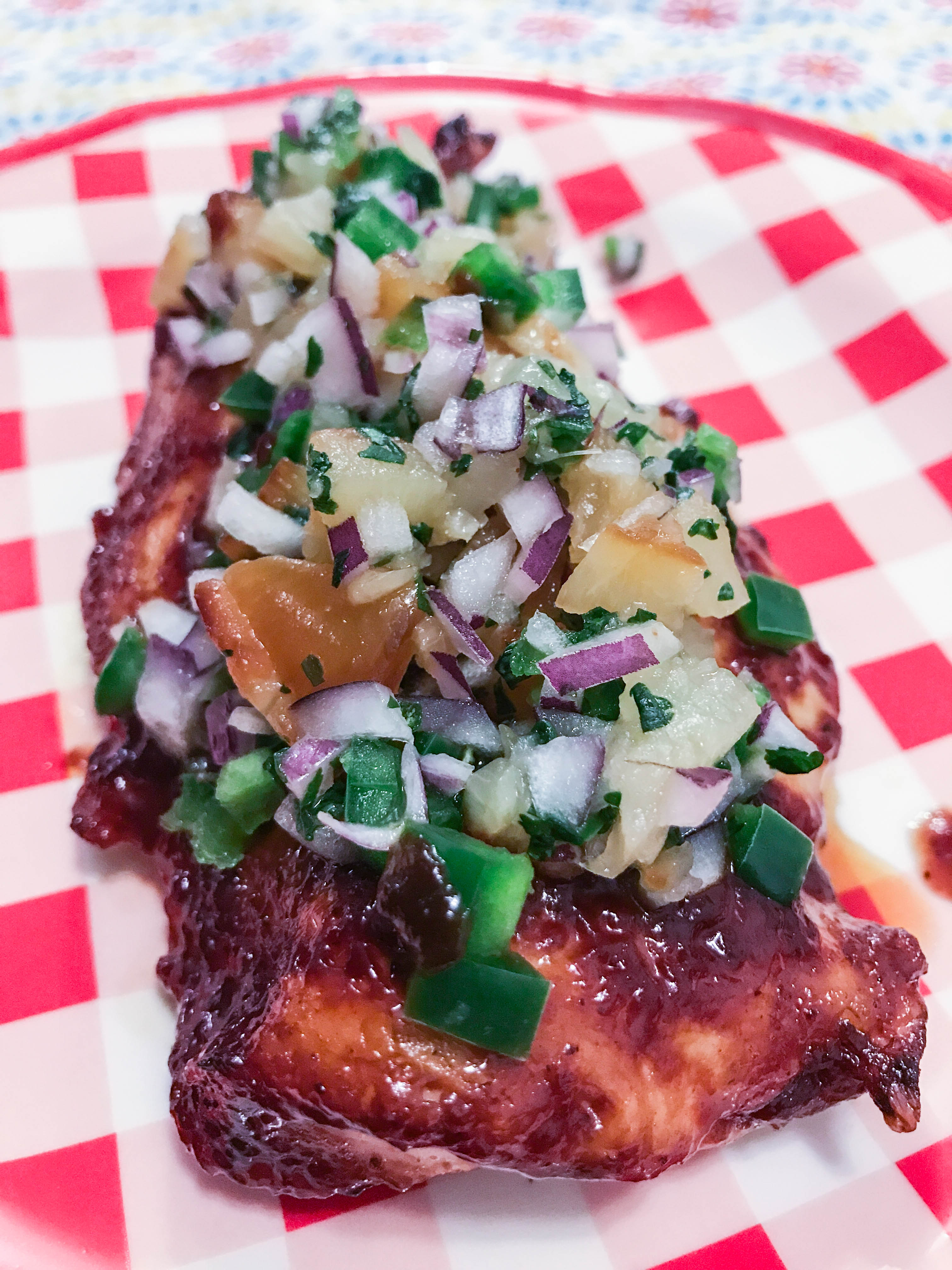 Honey jalapeno chicken breast topped with a big scoop of sweet pineapple salsa served on a red and white checked plate.
