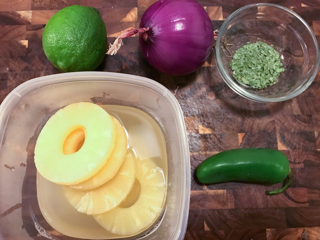 Slices of sweet pineapple, bright green lime, purple onion, and a spicy jalapeño on a wooden chopping block.