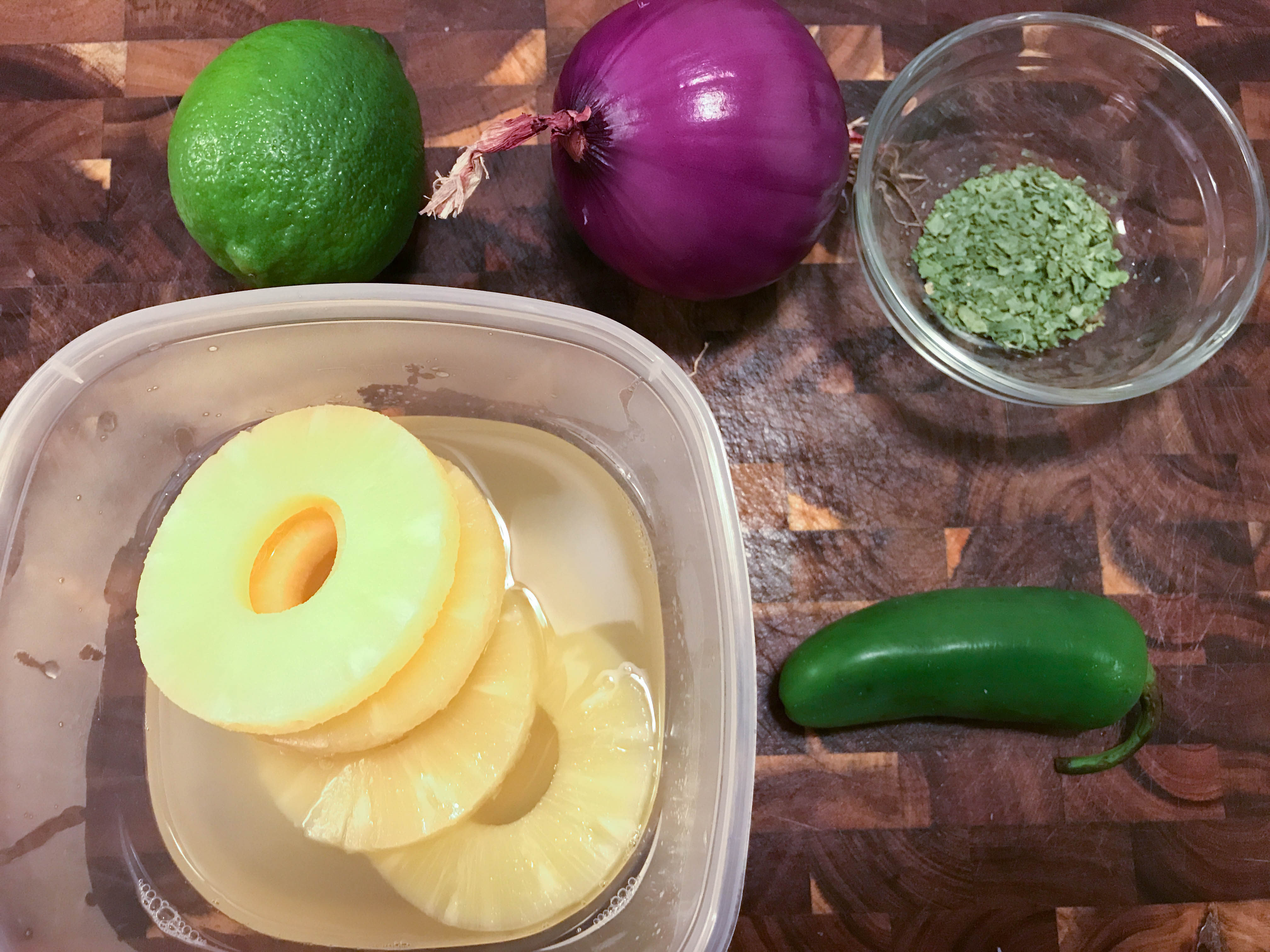 Slices of sweet pineapple, bright green lime, purple onion, and a spicy jalapeño on a wooden chopping block. 