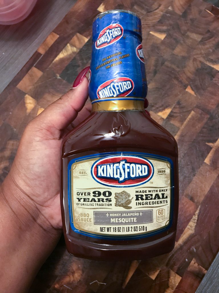 The secret ingredient (Kingsford Honey Jalapeño Mesquite BBQ sauce) being held above a wooden cutting board. 