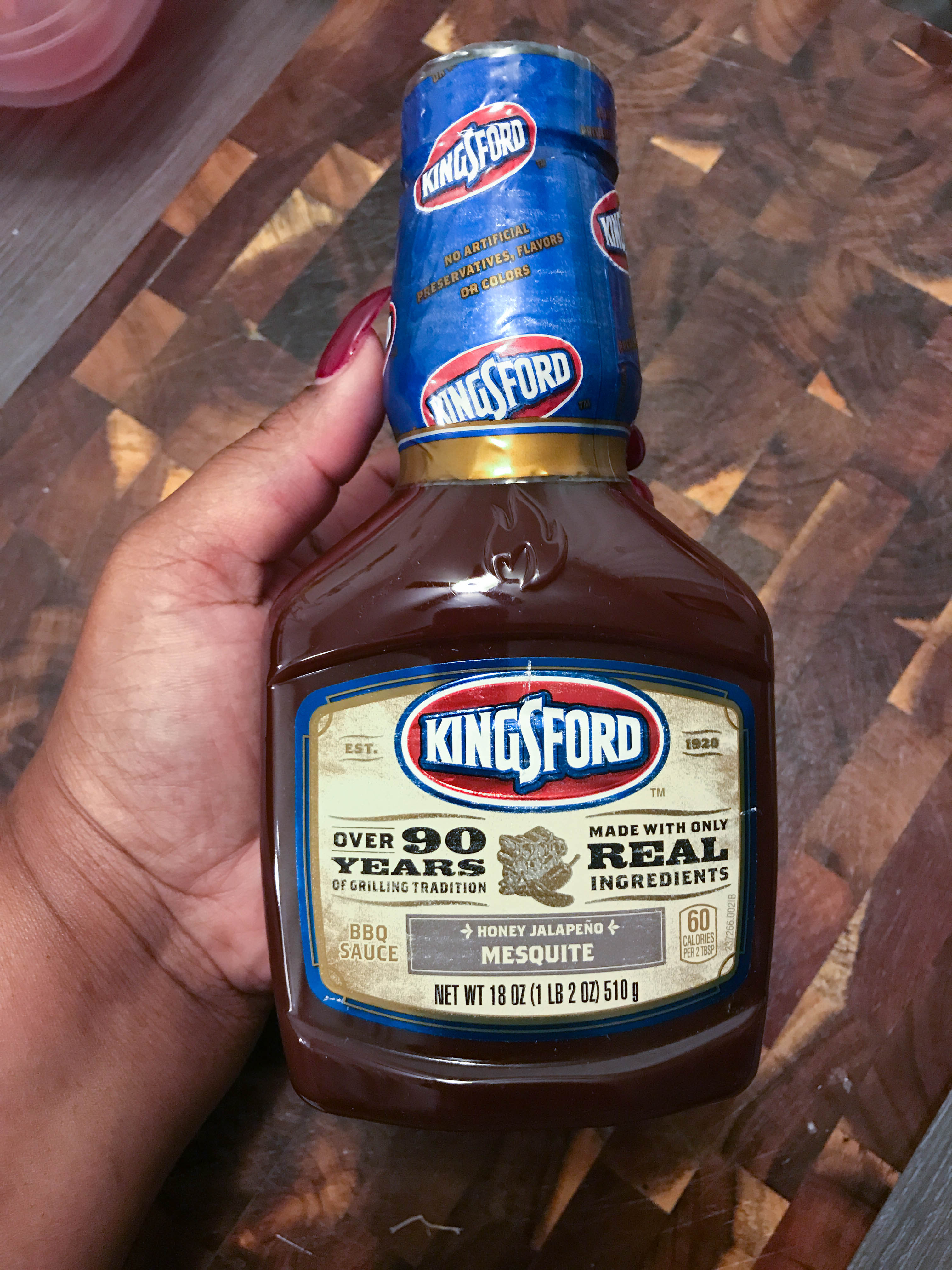 The secret ingredient (Kingsford Honey Jalapeño Mesquite BBQ sauce) being held above a wooden cutting board. 