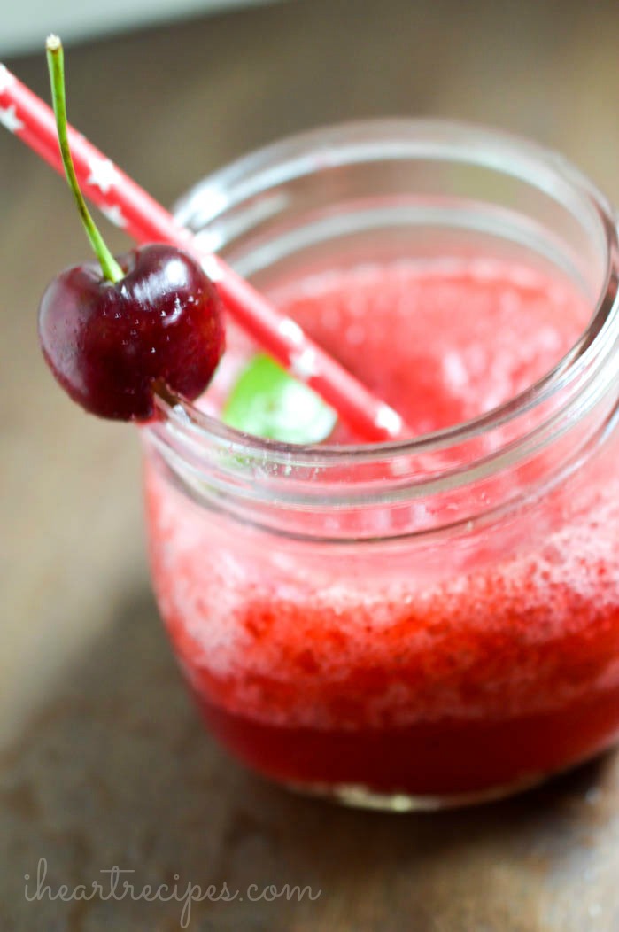 A sweet Cherry Lime Vodka Slushie in a frosty glass mason jar, served with a fun paper straw, and garnished with a red cherry. 