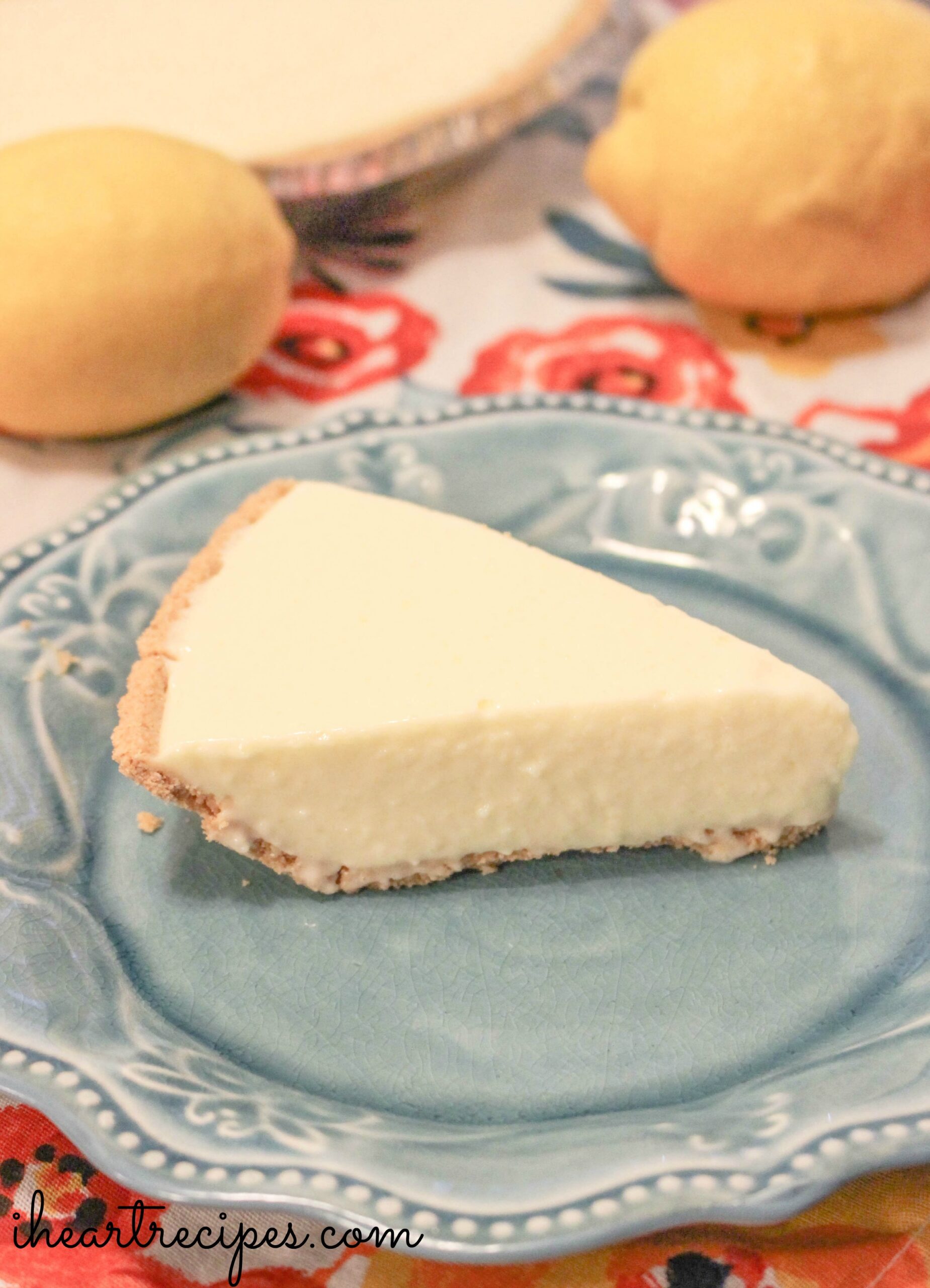 A single slice of no-bake lemon cheesecake served on a light blue ceramic dessert plate. In the background are two fresh lemons and the whole pan of lemon cheesecake served on the table.