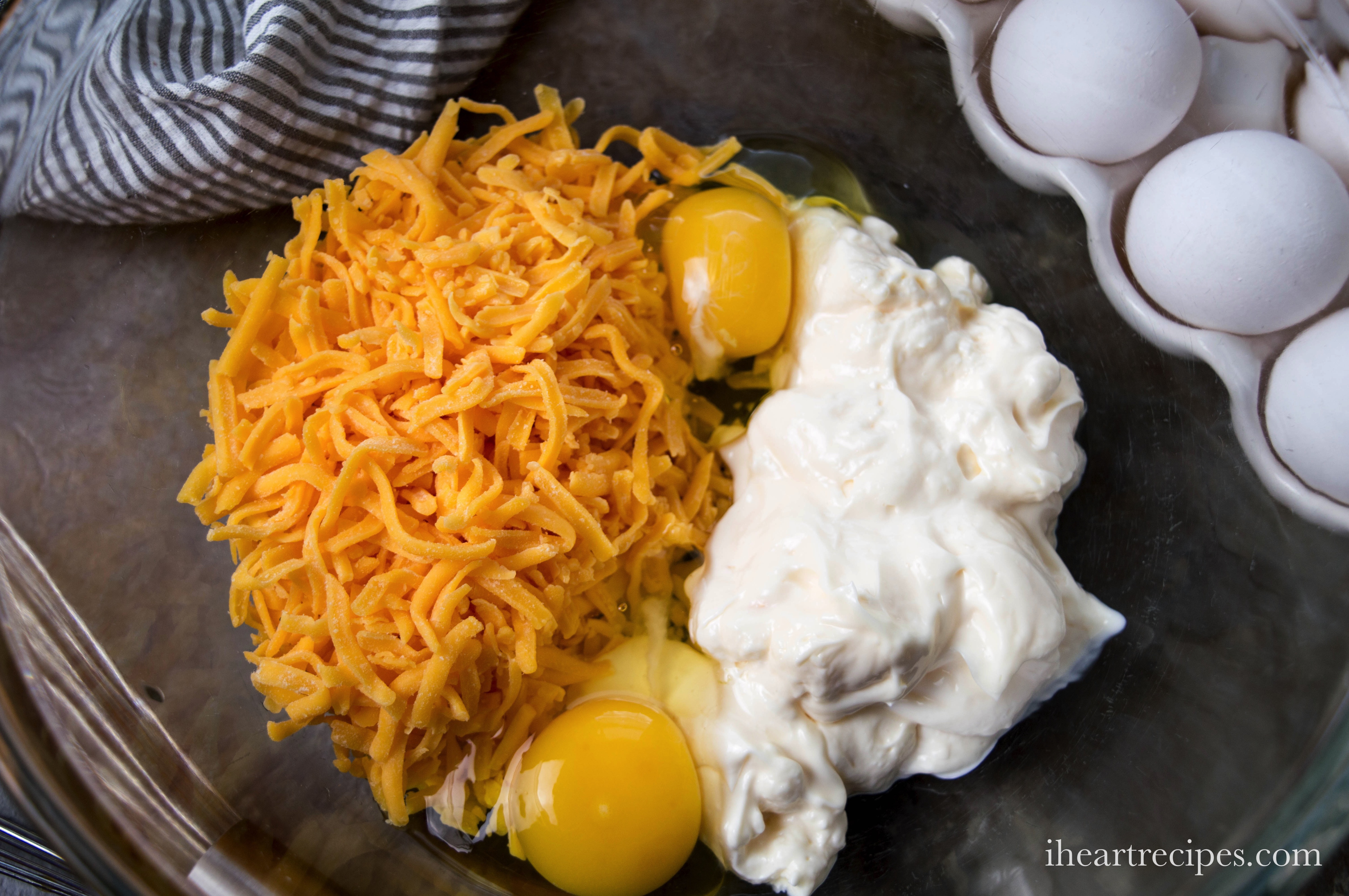 Mayonnaise, two eggs, and shredded cheese in a clear glass bowl. A carton of eggs sits next to the bowl on the brown countertop.