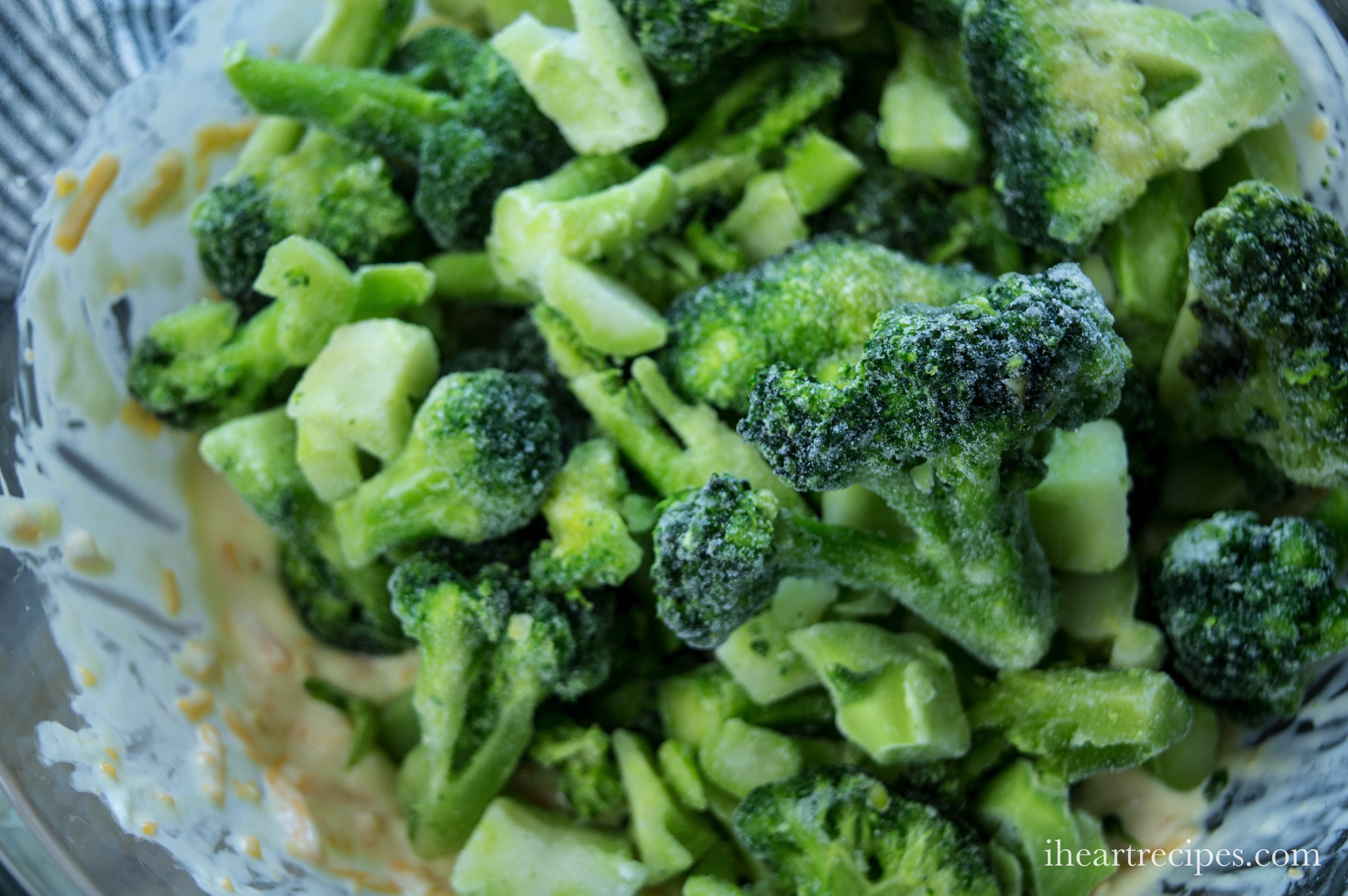 A pile of frozen broccoli florets sits in a bowl on top of a creamy mixture of mayonnaise and shredded cheese. This broccoli mixture is the body of a Southern-style broccoli casserole.