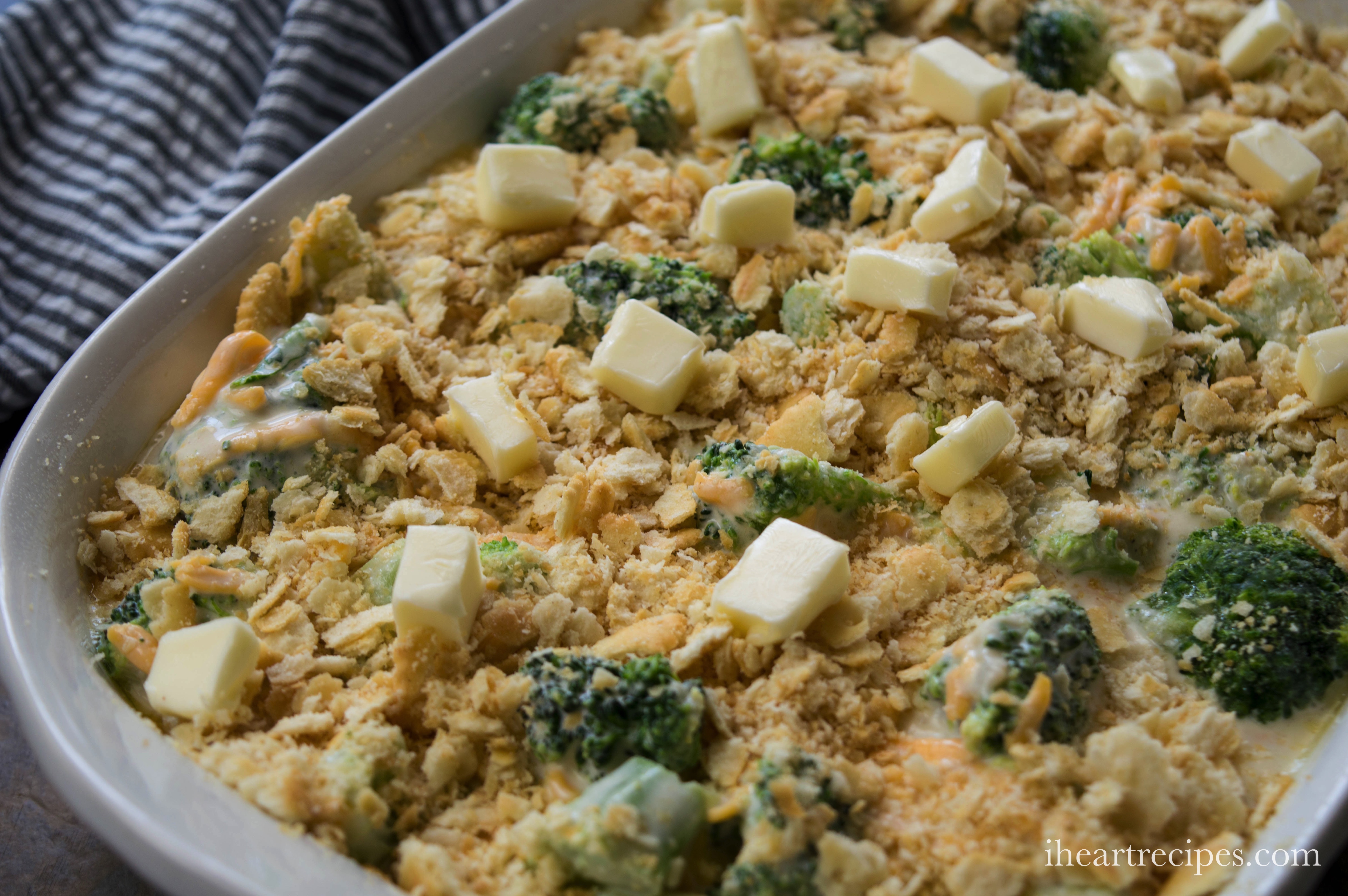 A white casserole dish is filled with an unbaked broccoli casserole. The top of the casserole is covered with crushed Ritz crackers and cubes of butter to make a crispy, buttery topping.