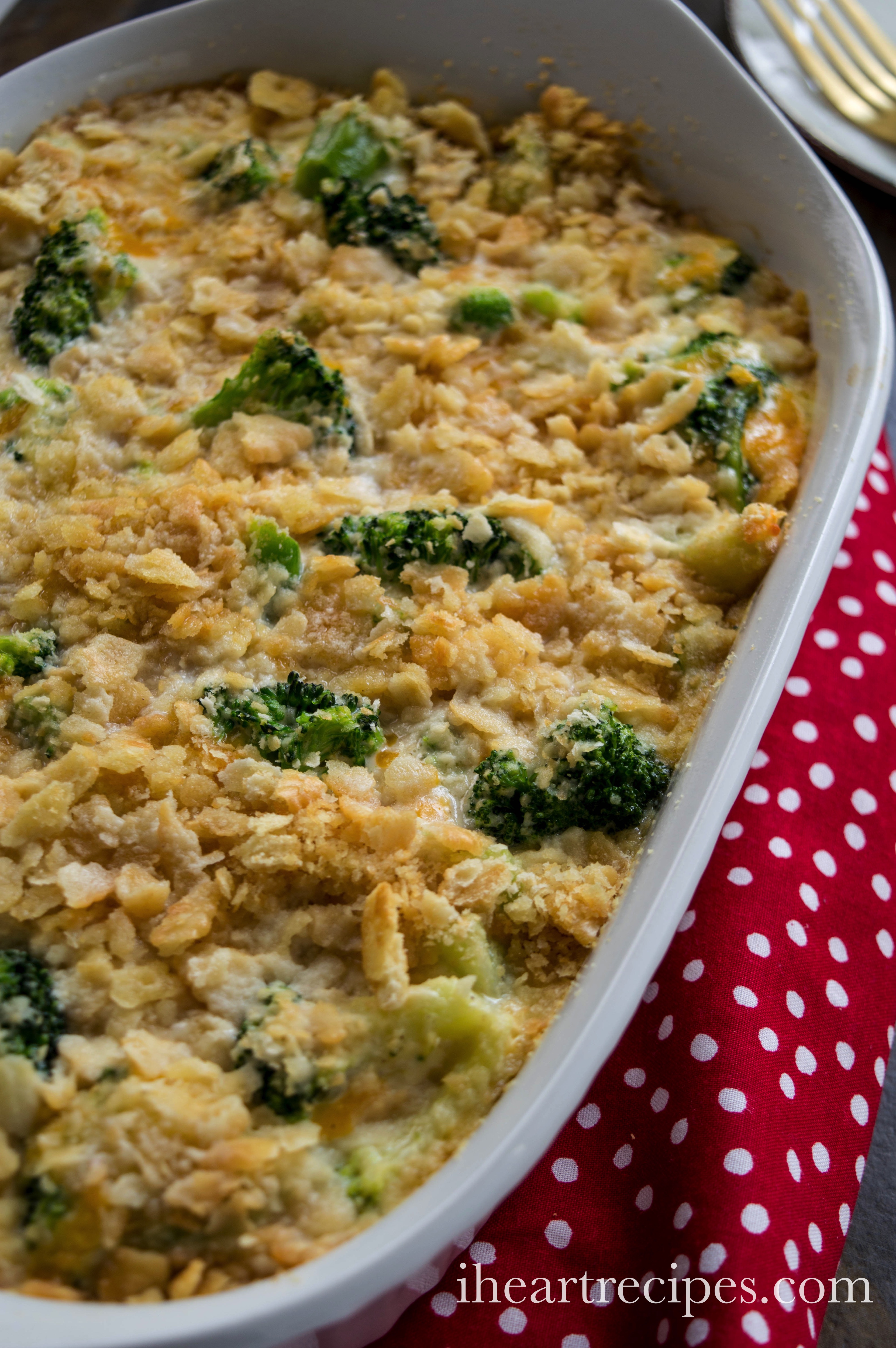 A freshly baked broccoli cheese casserole is served in a large white casserole dish, sitting on a red and white polka dot serving towel. The casserole has a crispy golden-brown cracker topping with florets of tender broccoli poking through.