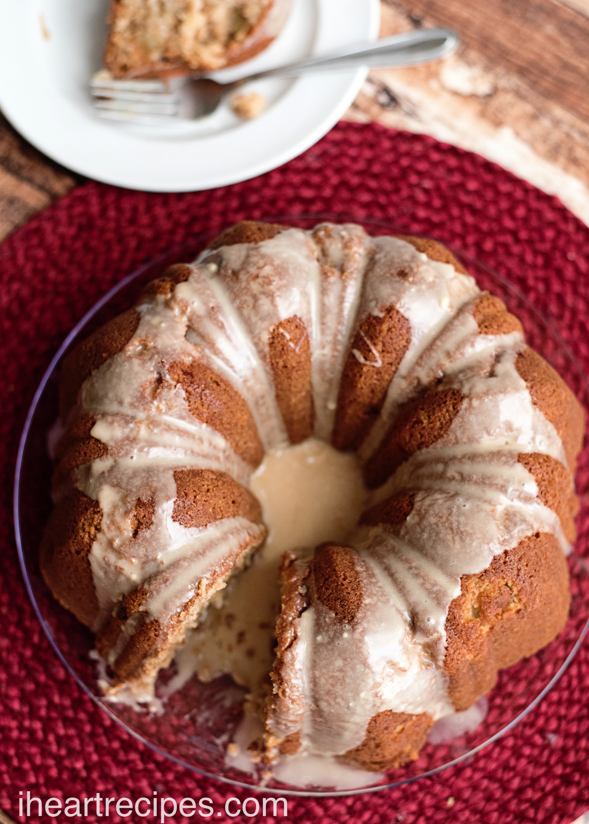 Fresh apple cake drizzled with maple glaze served on a clear glass platter on top of a red woven placemat. Try this recipe for a taste of fall sweetness!