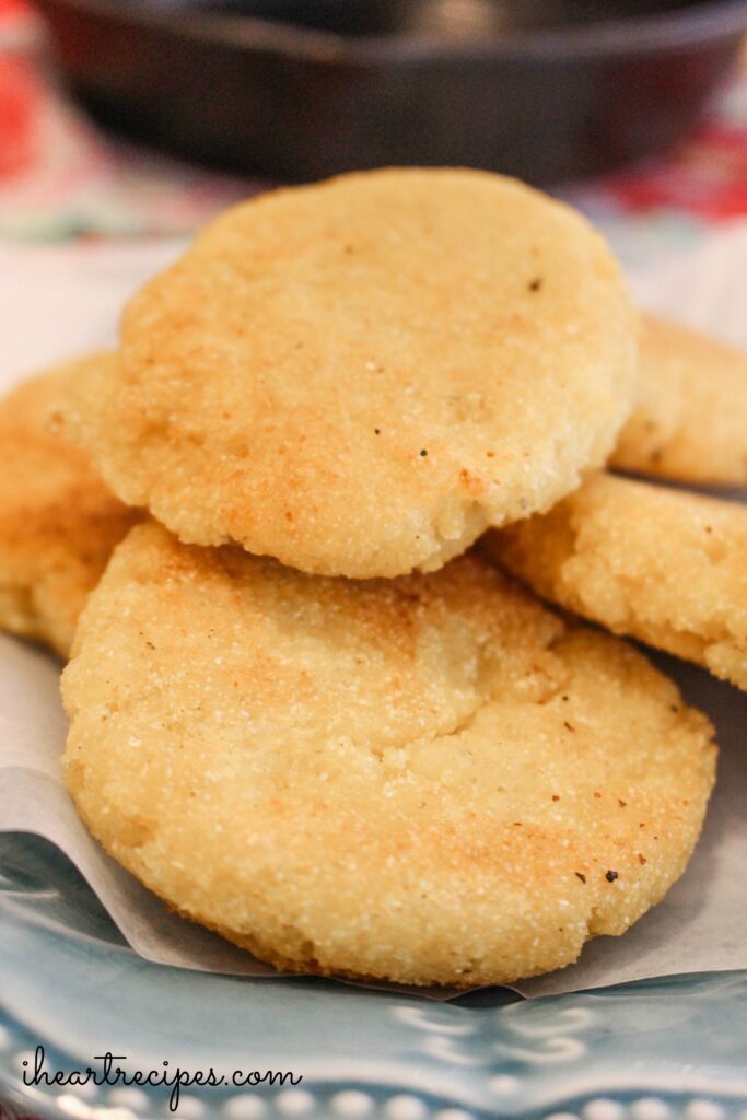 Five round pieces of hot water cornbread are stacked on a paper towel-lined plate. The cornbread is a perfect yellow-golden-brown color with a crispy exterior.