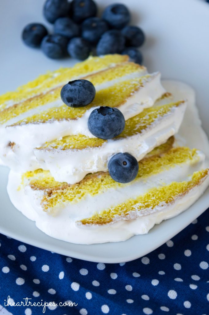 A closeup image of three slices of lemon ice cream cake. The creamy vanilla ice cream is melting into the layers of cake, topped with fresh blueberries as garnish.