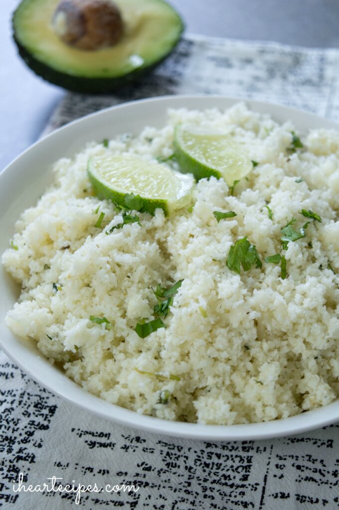 A white bowl filled with homemade riced cauliflower on top of a white and black fabric napkin. There is a halved avocado in the background. 