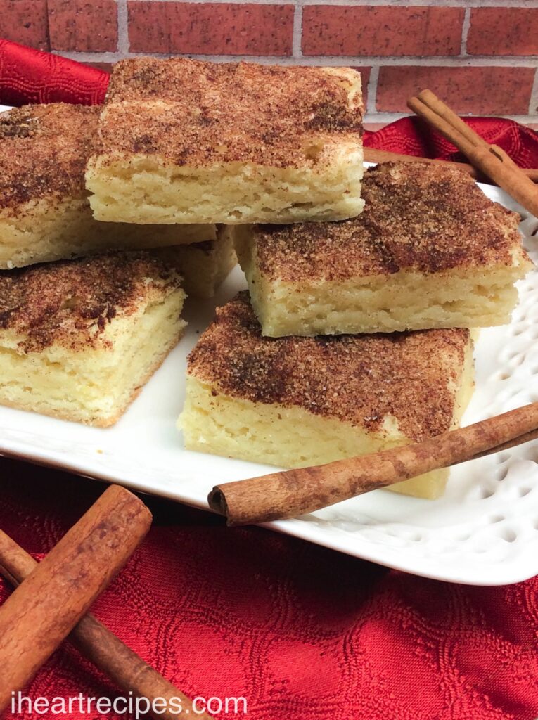 Moist and chewy Snickerdoodles bars stacked on a classy white platter and garnished with fragrant cinnamon sticks.